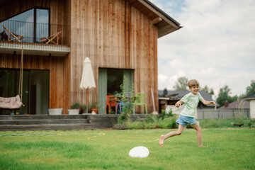 A young boy races across a grassy yard, laughing and having fun. His home, a modern wooden structure, stands in the background, surrounded by trees on a sunny day.