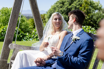Bride and groom sitting on swing, enjoying champagne and smiling outdoors