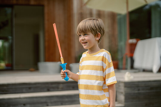 A young child stands in a backyard holding a blue toy sword with red handles, smiling happily. Sunlight floods the outdoor space, creating a joyful atmosphere.