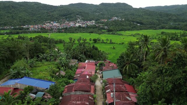A sky-high shot captures a rural village surrounded by lush forest, bright rice fields, and a distant town nestled at the base of tropical hills in Mahabang Tanaw, Quezon Province Philippines
