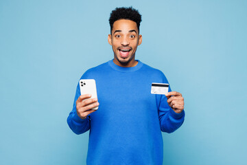 Young surprised man wear sweatshirt casual clothes using mobile cell phone hold credit bank card doing online shopping order delivery booking tour isolated on plain blue background. Lifestyle concept.
