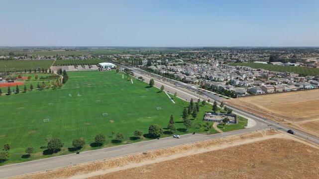 Side aerial shot starting over soccer and baseball fields, moving toward road with farmland and suburban houses in Ripon California.