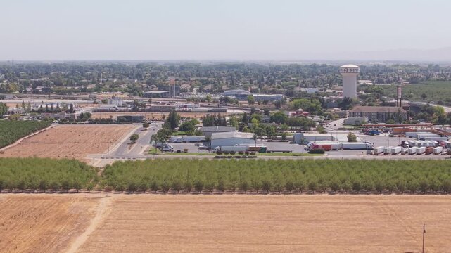 Side aerial shot along field with rows of trees, parking full of trucks, water tower, and busy road leading to Ripon California city.