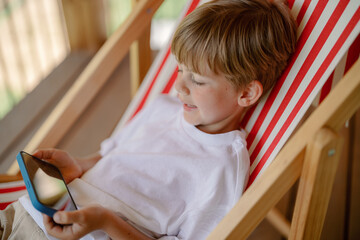 A young boy with short hair enjoys his time lounging in a striped deck chair, focused on a mobile device in his hands. Sunlight filters through the outdoor space, creating a serene atmosphere.