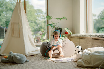 A young boy sits on a bean bag in a sunlit playroom, listening to a story on an electronic device while surrounded by toys, a tent, and bright green plants.