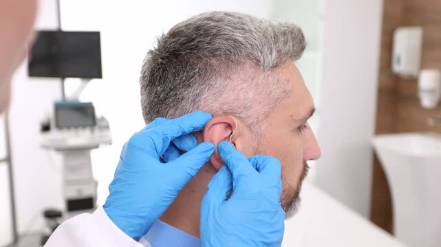 Installation hearing aid on man's patient ear at hearing clinic, close-up, side view. Deafness treatment, hearing solutions