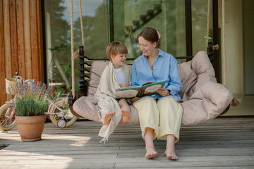 A mother reads a story to her child while sitting on a comfortable swing in their garden. It is a warm and sunny day, perfect for outdoor bonding time and imaginative adventures.