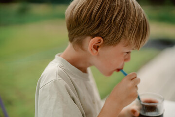 A young boy sips a cold beverage through a straw while outdoors. Bright sunlight highlights the green surroundings, creating a cheerful atmosphere perfect for relaxation.