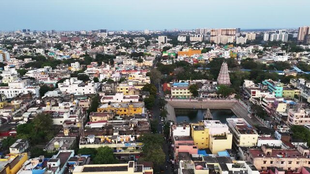 An aerial footage of a temple tank with a gopuram. The ancient temple landmark sits beside a street lined with modern, colorful buildings, showcasing the city's blend of old and new.