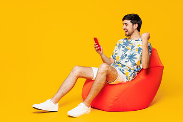 Full body young man wear blue beach t-shirt casual clothes sit in bag chair hold in hand use mobile cell phone do winner gesture isolated on plain yellow orange background studio. Lifestyle concept.