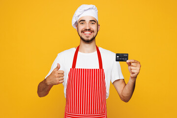 Young housewife housekeeper chef cook baker man wear red apron toque hat hold mock up of credit bank card show thumb up isolated on plain yellow orange background studio portrait Cooking food concept