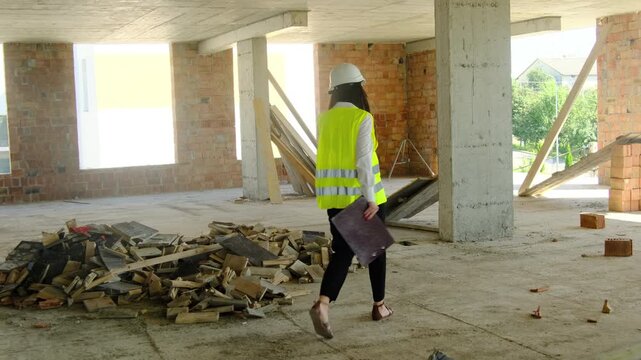 Lady inspector in hard hat and safety vest walks around work area. Engineer evaluates project stage and results of task completion
