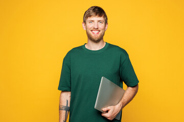 Young smart smiling cheerful happy IT man he wear green t-shirt casual clothes hold closed laptop pc computer look camera isolated on plain yellow orange background studio portrait. Lifestyle concept.
