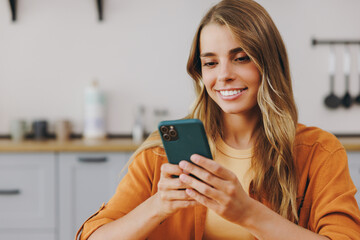 Close up young smiling happy housewife woman wear orange shirt casual clothes hold in hand use mobile cell phone chat online sit at table in light kitchen at home alone Lifestyle cooking food concept