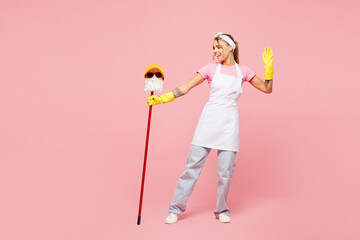 Full body young woman wear white apron gloves hold funny mop in glasses and hat pov human man while doing housework tidy up dance isolated on plain pastel light pink background. Housekeeping concept.