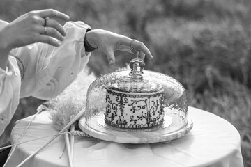 Artistic black and white photo of elegant hands with a cake under a glass dome, symbolizing sophistication, celebration and timeless style