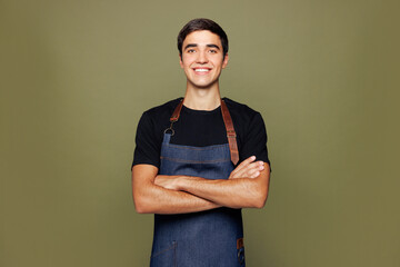 Young man barista bartender barman employee wear jeans apron black t-shirt work in coffee shop hold hands crossed folded look camera isolated on plain green background. Small business startup concept.