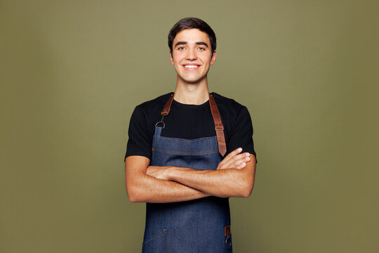Young man barista bartender barman employee wear jeans apron black t-shirt work in coffee shop hold hands crossed folded look camera isolated on plain green background. Small business startup concept.