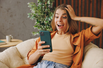 Young surprised woman wear orange casual clothes hold in hand use mobile cell phone sits in chair stay at home hotel flat rest relax spend free spare time in living room indoor. People lounge concept.