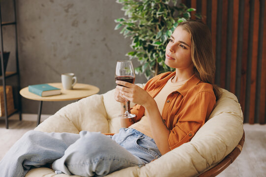 Side profile view young minded calm woman wear orange casual clothes drink red wine sits in chair stay at home hotel flat rest relax spend free spare time in living room indoor. People lounge concept.