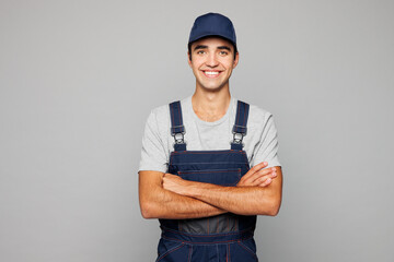 Young happy confident car mechanic man he wear denim overall hat uniform workwear clothes work in garage look camera isolated on plain grey background studio portrait. Automotive repair job concept.