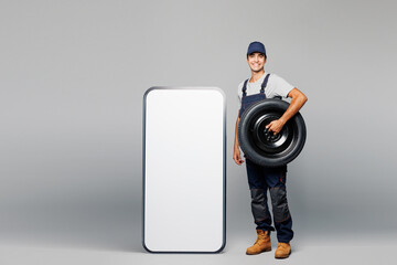 Full body young car mechanic man in overall uniform workwear clothes work in garage big huge blank screen mobile cell phone hold wheel isolated on plain grey background Automotive repair job concept