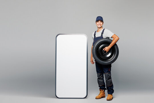 Full body young car mechanic man in overall uniform workwear clothes work in garage big huge blank screen mobile cell phone hold wheel isolated on plain grey background Automotive repair job concept