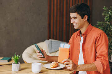 Traveler young man wear casual clothes hold passport ticket sit at table in coffee shop cafe relax rest in restaurant indoors. Tourist travel abroad in free time rest getaway. Air flight trip concept.