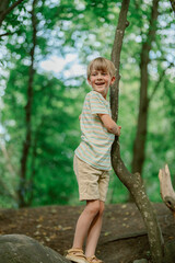 Naklejka premium A young boy stands joyfully on a rock, holding onto a tree trunk. Surrounded by lush green foliage, he enjoys a playful moment in the serene forest environment.