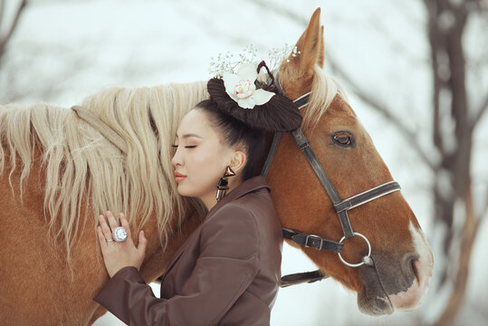 Elegant asian woman in vintage hat embracing horse in winter nature. Almaty, Kazakhstan.