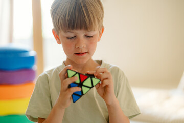 A young boy concentrates on solving a colorful triangular puzzle in a bright indoor space. He is focused and engaged, surrounded by vibrant cushions in the background.