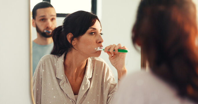 Couple, brushing teeth and mirror in bathroom for dental hygiene or morning routine in home. Man, woman and toothbrush with reflection for fresh breath, clean mouth or oral care together in house - Powered by Adobe