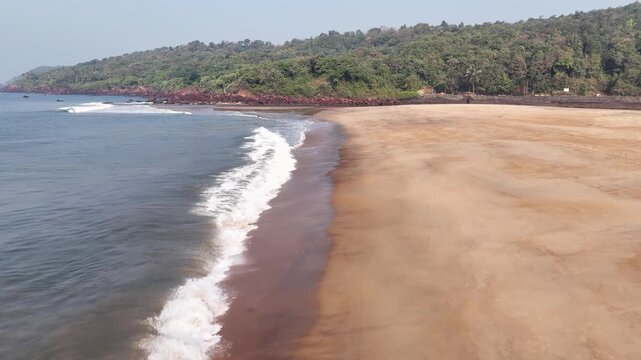 Low-altitude drone shot flying over an empty tropical beach, showing waves washing onto the golden sand at Kelus Kalvi, India.