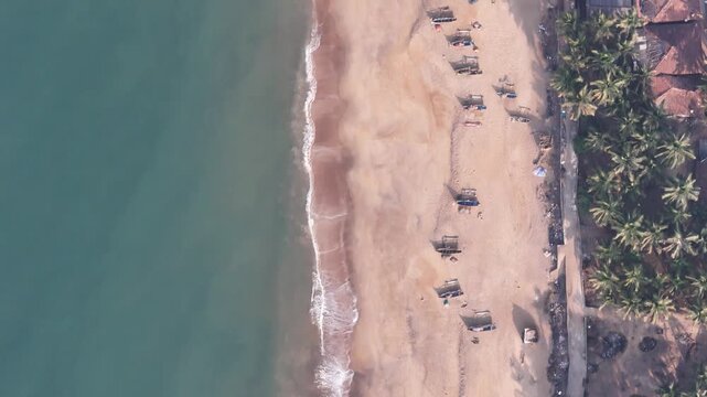 Top-down aerial view of a busy fishing beach in India, with fishermen and traditional boats on the sand as waves wash ashore.