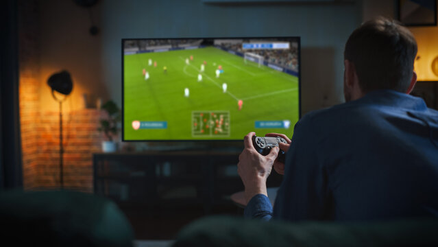 Young Couple Having an Exciting Evening at Home, Sitting Together on a Sofa and Playing a Soccer Video Game on a Console. Girlfriend Supporting Her Gamer Boyfriend, Watching Action Unfold on TV - Powered by Adobe