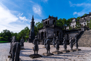 A wide view of the Khai Dinh Tomb's courtyard, featuring rows of stone statues and an ornate, dark monument under a bright blue sky.