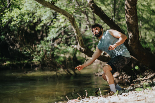 A young man in casual attire skips stones by a peaceful stream surrounded by forest vegetation, enjoying a relaxed outdoor activity.
