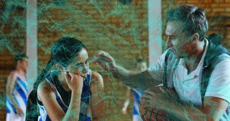 Leaning coach holding basketball, speaking to teen player on gym sideline bench, with team jerseys