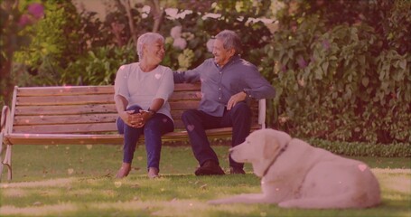 Sitting older couple smiling at each other on wooden bench in garden, with light-colored dog