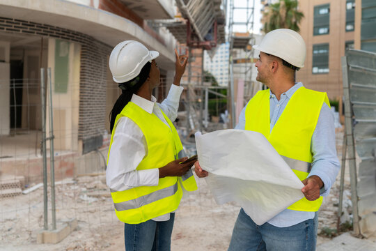 Two diverse Architects in hardhats and safety vests examining construction plans on a building site
