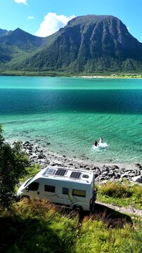 Two travelers embrace the clear turquoise waters of the Lofoten Norway, with a modern camper van parked on the serene shoreline. The beautiful landscape features majestic mountains 