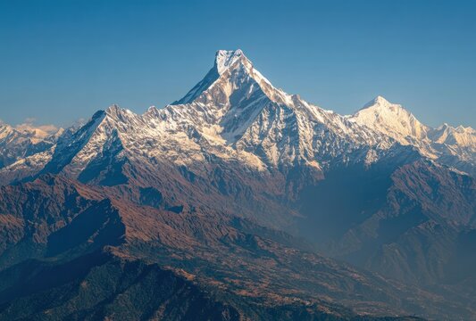 A majestic mountain peak, snow-capped and rugged, rises above a vast landscape of forested hills under a clear, blue sky