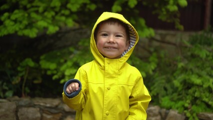 child in a yellow raincoat with hood smiles in the rain with green garden background