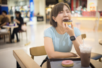 Woman wearing a blue t-shirt is eating a donut while sitting at a donut shop