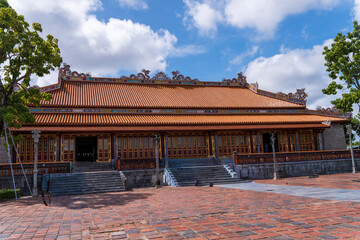 A traditional royal building with an orange tiled roof and detailed decorations stands on a vast brick courtyard in Hue, under a bright, cloudy sky.