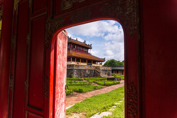 A stunning view through a vibrant red doorway reveals a traditional palace with an orange roof and stone base, surrounded by manicured gardens under a cloudy sky.