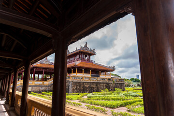 A tranquil garden courtyard with traditional buildings and manicured hedges is viewed through the dark wooden corridor of an ancient palace in Hue under a cloudy sky.