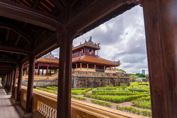 A tranquil garden courtyard with traditional buildings and manicured hedges is viewed through the dark wooden corridor of an ancient palace in Hue under a cloudy sky.
