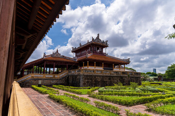 A tranquil garden courtyard with traditional buildings and manicured hedges is viewed through the dark wooden corridor of an ancient palace in Hue under a cloudy sky.