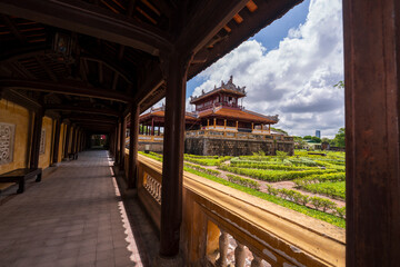 A tranquil garden courtyard with traditional buildings and manicured hedges is viewed through the dark wooden corridor of an ancient palace in Hue under a cloudy sky.